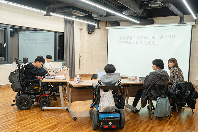 좌담회 전경. V자 대형으로 앉아 있는 좌담 참여자들의 뒷모습과 옆모습.
좌담 참여자 홍성훈이 노트북 키보드 위에 오른손을 올려 텍스트를 작성하는 중이고,
앞쪽 벽면에 설치된 커다란 스크린에 그 텍스트가 실시간으로 영사된다.
스크린에는 “일단 황철호 배우님의 스타일리시한 휠체어와 외모가 인상적이”까지 문장이 쓰여 있다.