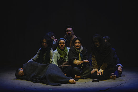 <화전>의 공연 사진. 어두운 무대에 푸른색의 조명이 한 무리의 인물들을 비춘다. 이들은 모두 목도리, 담요 등을 두르고 바닥에 앉거나 누워 추위를 견뎌내고 있다. 