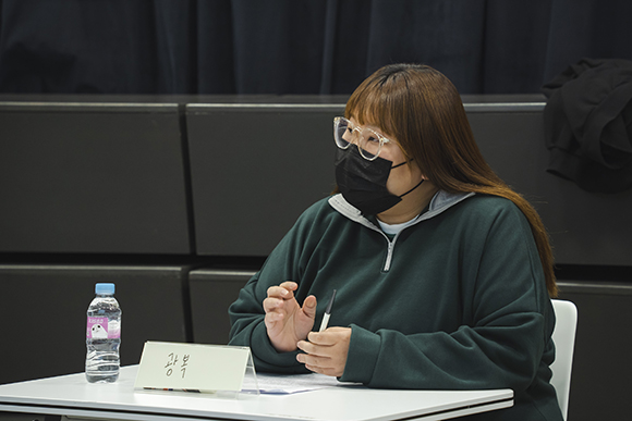 작가 구지수. 진한 초록색의 티를 입고, 투명한 뿔테 안경을 착용했다. 밝은 갈색으로 염색한 긴 생머리를 늘어뜨렸다. 책상 위에 놓인 이름표에는 ‘광복’이라고 쓰여 있다. 