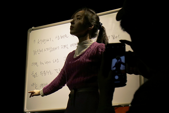 <연극 안 하기 1 - 단단히 경고하기>의 공연 사진. 분홍색 니트에 흰 폴라티를 받쳐 입은 전혜인이 오른손을 가볍게 뻗고 고개를 쳐들어 발레 동작을 취하고 있다. 전혜인의 뒤에는 흰 화이트보드가 있다. 화이트보드에는 “서연씨, 상체를… 자신이 세상에서 제일… 예쁜 척해야 돼요. 좋은 무용수는 결국…” 등이 쓰여 있다. 휴대폰 카메라로 전혜인과 화이트보드를 촬영하는 사람의 실루엣과 휴대폰 화면이 보인다.
