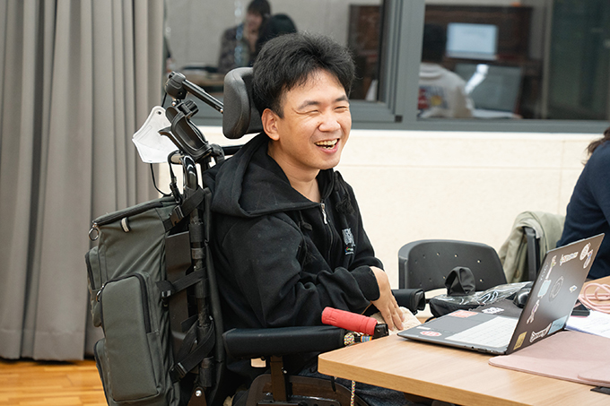 좌담 참여자 홍성훈. 검은색 후드 짚업을 입고 있다. 건너편을 바라보며 환하게 웃는 얼굴이다.
등받이에 카키색의 큰 배낭이 걸려 있는 전동휠체어에 앉아 있고, 책상 위에는 노트북을 펼쳐두고 있다.