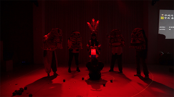 <안타 오이디푸스: hit or run>의 공연 사진. 붉은 조명이 무대 전반을 밝히고 있다. 실루엣만 보이는 한 명의 배우가 무대 중앙에 무릎을 꿇고 손을 등 뒤로 모아 고개를 숙이고 있고, 그의 뒤로 다섯 명의 배우가 그를 둘러싸고 서 있다. 다섯 명의 배우 중 중앙의 배우는 머리에 희고 큰 깃털 세 개를 꽂고 청사초롱을 두 손으로 들고 서 있다. 나머지 네 명의 배우는 상체와 머리를 모두 가리는 커다란 가면을 두 손으로 들고 있는데, 가면은 각각 다른 얼굴을 묘사한 것으로 고대의 벽사가면 같은 분위기를 풍긴다.