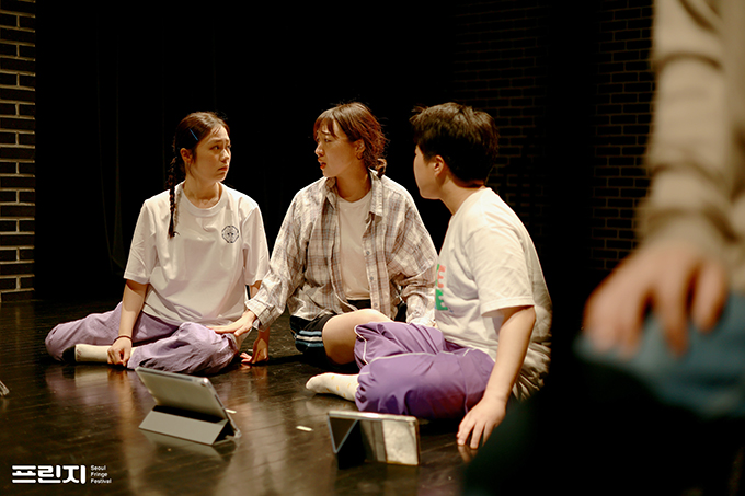 <홀리섹스데이> 공연 사진. 다영, 해지, 다빈이 바닥에 모여 앉아 심각한 표정으로 이야기를 나누고 있다. 이들은 모두 트레이닝 팬츠와 흰 티셔츠를 입은 편안한 차림이다. 
            세 사람의 앞에 태블릿PC 두 대가 놓여 있고, 사진의 우측으로는 의자에 앉은 누군가의 무릎과 그 위에 올린 손이 흐릿하게 보인다. 