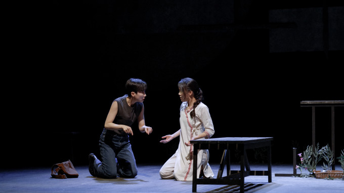 <맆소녀> 공연 사진. 연영과 까이가 나란히 무릎을 세워 앉은 자세로 서로의 눈을 보고 있다. 짧은 커트 머리를 하고 검은 민소매 티셔츠에 청바지를 입은 연영은 양손의 검지를 펴서 바닥을 가리키고, 머리를 길게 땋아 묶고 가장자리를 따라 붉은색 나뭇잎이 수놓아진 하얀 옷을 입은 까이는 양손의 손바닥을 위로 향하게 하고 있다. 그 옆에 검은색 나무 벤치와 키가 작은 식물들이 있다.