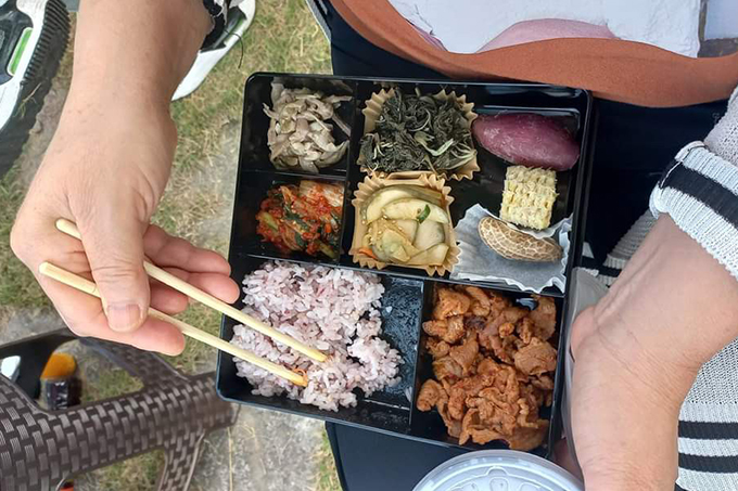 잔디밭을 배경으로 나물과 밥, 돼지불고기, 김치, 고구마 등이 담긴 도시락이 한 사람의 무릎에 놓여있다.
사진 속 손은 젓가락으로 밥을 뜨고 있다.