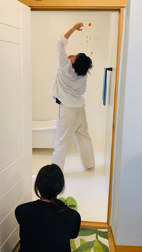 <맡는 경계>의 전시 연계 퍼포먼스 사진. 공간의 열린 문 너머로 흰 셔츠와 옅은 베이지색 바지를 입은 곽혜은 작가가 등을 보인 채 왼손을 높이 들고 움직이는 것이 보인다. 
            고개는 한껏 뒤로 젖혀 감은 두 눈이 보이고, 위로 올린 왼손은 무엇을 쥔 모양으로 구부러져 있다. 
            곽혜은의 뒤로 욕조의 일부와 파란 수건이 걸린 수건걸이가 보인다. 문 안쪽에 연두색 욕실화가 놓여 있다. 
            문 밖에는 검은 옷을 입은 한 사람이 초록색 나뭇잎이 들어간 베이지색 러그에 앉아 곽혜은을 바라본다.