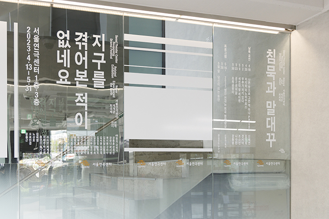 서울연극센터 유리벽에 희곡제 <침묵과 말대꾸>, 전시 <지구를 겪어본 적이 없네요>의 개요가 흰색 텍스트로 세로쓰기 되어 있다. 행사 일정과 참여 작가의 이름들이 쓰여 있고, 유리에 반사된 공간 내외부와 유리 너머에 있는 공간 계단 등이 겹쳐 보인다.