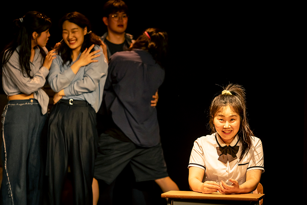 <고등어>의 공연 사진. 교복을 입고 머리를 높게 하나로 묶은 지호가 책상에 앉아 들뜬 표정으로 정면을 응시한다. 그의 뒤편으로 존재들이 웃음 짓거나 서로를 부둥킨 채 서 있다.
