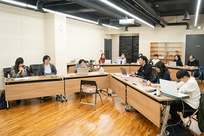 좌담회 전경. 좌담 참여자 하은빈, 임대륜, 좌담 진행자 김지수, 좌담 참여자 홍성훈, 양근애, 임지윤이
테이블 네 개를 각도가 넓은 V자 형태로 붙인 채 앉아 있다.
그 뒤쪽으로 참관인들이 책상 세 개를 붙여 앉아 있다. 모두 살짝 웃고 있는 얼굴이다.