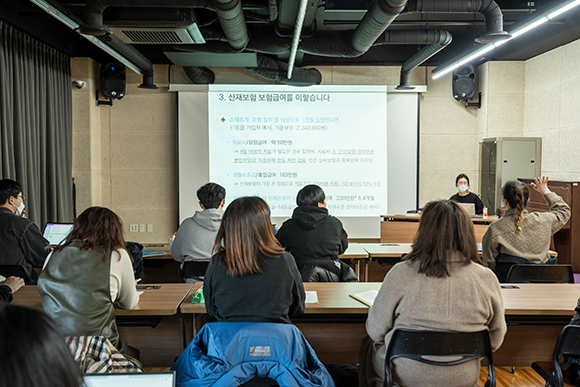 ‘예술인 산재보험 찾아가는 설명회’ 현장 사진. 벽면 스크린에 산재보험 보험급여에 대한 사례가 제시된 프리젠테이션 화면이 띄워져 있다. 그 옆으로 설명회를 진행한 한국예술인복지재단 사회보험팀 남희승 팀장이 앉아 있다. 맞은편에는 열을 맞춰 책상이 놓여 있고, 각자 노트북이나 노트에 메모를 하고 있는 참여자들의 뒷모습이 보인다. 참여자들 중 한 사람이 손을 들어 질문하고 있다.