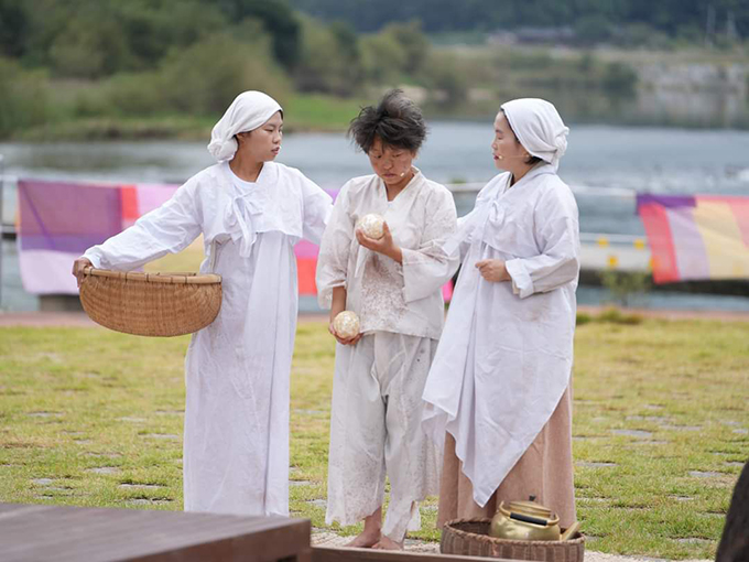 <심청길 비밀레시피>의 공연 사진. 흰 저고리와 흰 치마, 흰 머릿수건을 하고 옆구리에 광주리를 낀 사람과, 흰 저고리와 흰 머릿수건,
흰 앞치마, 베이지색 하의를 입은 사람이 가운데 선 사람의 등을 도닥이고 있다.
가운데 선 이는 더벅머리를 하고, 때가 묻은 흰색 상하의를 입었다. 맨발에 바지 밑단이 찢어져 있으며, 양손에 각각 주먹밥 한 덩이씩을 들고 있다.
세 사람의 뒤로 푸른 잔디밭에 색색의 천이 빨래줄에 널려 있고, 강물이 흐르고 있다.