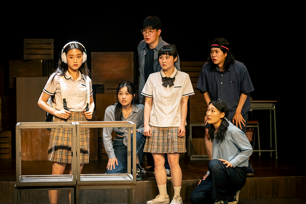 <고등어>의 공연 사진. 정육면체 모양의 철제 프레임 두 개를 이어 붙여 만든 수조 앞에 헤드폰을 끼고 흰 백팩의 어깨끈을 가볍게 쥔 채 허공을 바라보는 경주가 있다. 그의 뒤편으로 경계심 넘치는 표정의 지호와 호기심 넘치는 표정의 존재들이 경주를 바라본다. 경주와 지호는 교복차림이고, 존재들은 각자 짙고 연한 파란색의 사복을 입었다. 