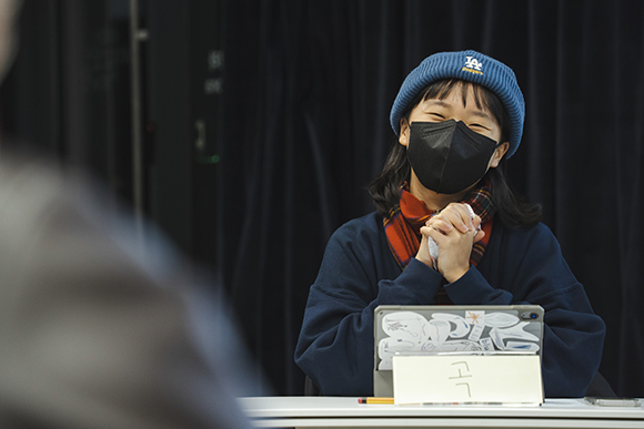 작가 김영화. 파란 LA 다저스 비니를 쓰고, 짙은 남색의 맨투맨과 빨간 체크무늬 목도리를 두르고 있다. 두 손으로 핫팩을 쥐고, 웃고 있다. 