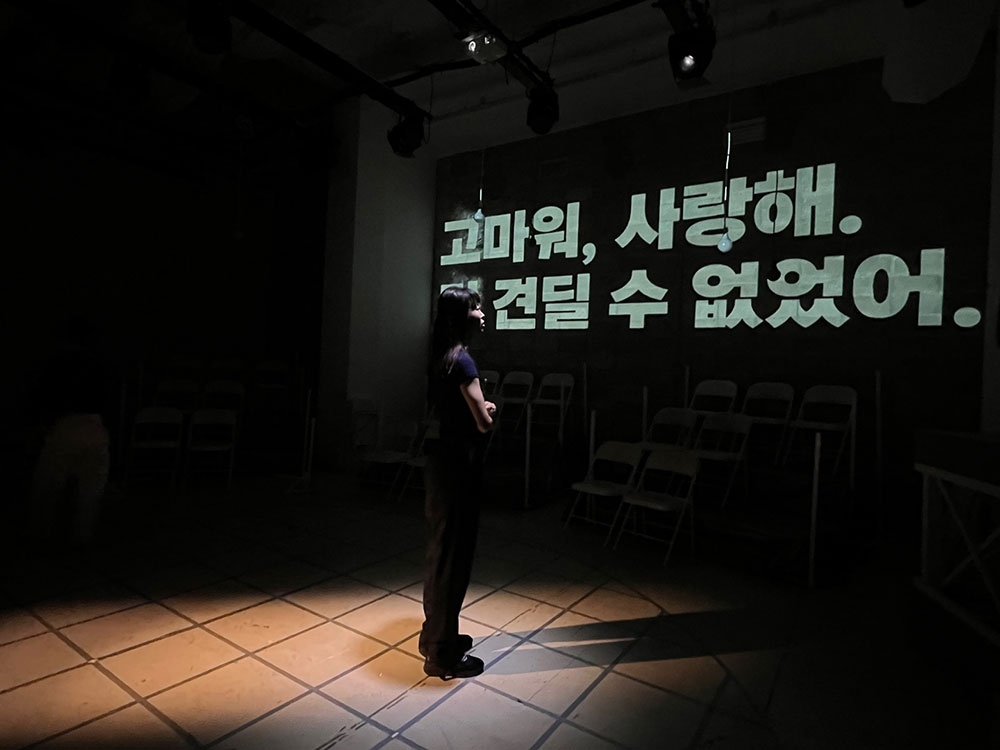 <캐빈방정식>의 공연 사진. 벽면에 커다란 글자로 ‘고마워, 사랑해. 더 견딜 수 없었어.’라는 문구가 커다랗게 영사된다. 한 인물이 이를 바라보며 팔짱을 끼고 서있다.