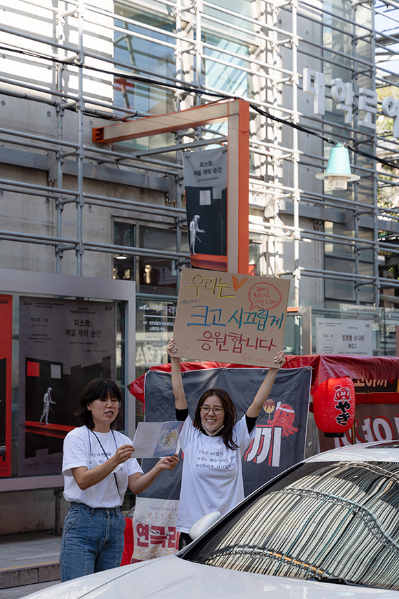 대학로예술극장 앞에서 ‘미투 이후 다시 말하기’ 퍼포먼스가 진행되고 있다. ‘우리는 크고 시끄럽게 응원합니다’라는 피켓을 머리 위로 번쩍 든 여성이 활짝 웃고 있고 그 옆에서 소리 내어 무언가를 읽고 있는 여성의 모습도 보인다. 퍼포머들의 뒤편에는 다코야키를 파는 노점이 보이고, 흰 승용차가 이들의 앞을 지나가고 있다.