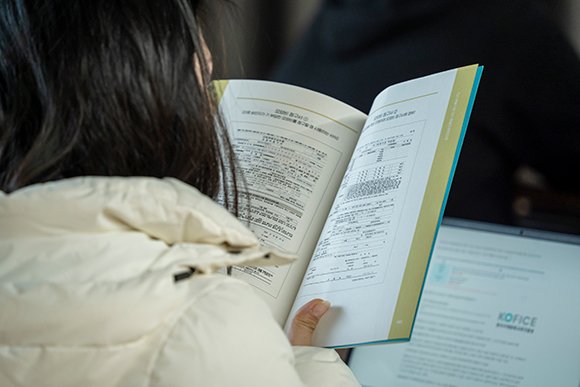 ‘예술인 산재보험 찾아가는 설명회’ 현장 사진. 한 참여자가 설명회에서 제공된 『예술인 산재보험 혜택이야기』 책자를 펼쳐 읽고 있다. 어깨 너머로 ‘요양비 청구서’ 신청양식이 보인다.