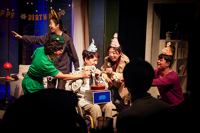 <생일파티> 공연 사진. 민수와 희정, 엄마와 경찰이 함께 모여 생일파티를 하고 있다.
민수 역의 배우는 경찰 제복을 입고 머리에 고깔을 쓴 채 무릎 위에 케이크를 올려 놓았다.
엄마 역의 배우는 초록색 원피스에 망사와 리본, 구슬 등으로 만들어진 초록색 머리 장식을 얹었다.
경찰 역의 배우는 레이스가 달린 자줏빛의 내복을 입은 채다.
그들의 뒤쪽으로 도둑 역할의 배우가 옆쪽을 바라보며 한 손을 쭉 뻗어 반갑게 누군가를 맞이하고 있다.