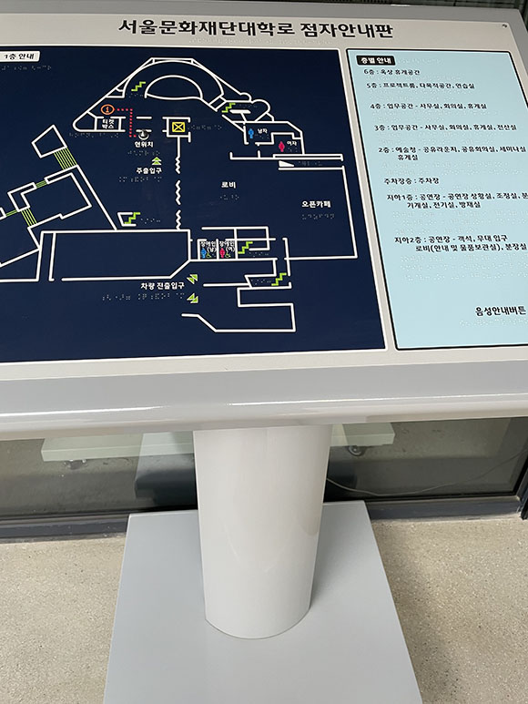서울문화재단대학로 청사의 점자 안내판을 촬영한 사진이다. 안내도 좌측에는 건물의 1층 촉지도가 있으며, 오른쪽에는 1층부터 6층까지의 공간에 대한 층별 정보가 점자와 묵자로 안내되어 있다.