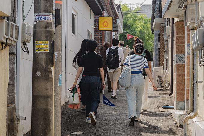 <오프 리밋> 현장 사진. 관객들이 안정리 골목길을 따라 기지촌여성평화박물관 일곱집매로 찾아가고 있는 모습을 뒤에서 찍은 사진이다. 
            가장 앞쪽에 군복을 입고 깃발을 든 채 관객들을 안내하는 배우가 보인다. 
            노란색 바탕에 빨간색 꽃이 그려진 일곱집매의 작은 간판이 건물 벽에 걸려 있다.