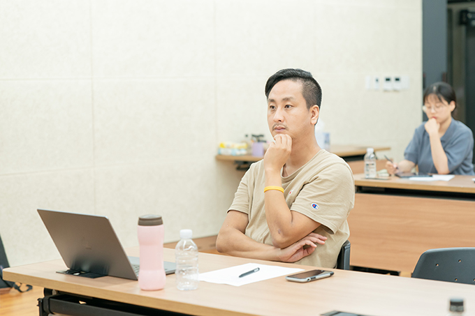 연출가 전윤환. 베이지색 반소매 티셔츠를 입고 투 블록 컷의 머리를 한쪽으로 넘겼다.
왼팔에 노란 밴드의 팔찌를 하고 있으며, 손을 턱에 얹은 채 정면을 바라보고 있다.