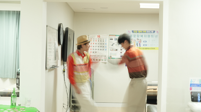 <재개발 옆 경로당>의 공연 사진. 흰 벽으로 둘러싸인 공간의 벽에는 화이트보드, 시계, TV, 달력, 경로당 복지 관련 현수막 등이 걸려 있다.
그 가운데서 두 남성이 마주 보고 춤을 추고 있다. 사진상 이들의 팔과 다리가 흐리게 드러나 그 움직임을 짐작할 수 있다.