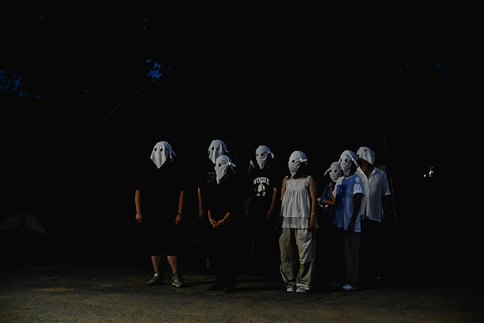 <정전의 밤> 공연 사진. 얼굴에 하얀 천을 덮어 쓴 여덟 명의 사람이 야외에 서 있다. 
            흰 천에는 두 개의 구멍이 뚫려 앞을 볼 수 있으며, 사람들은 귀 옆으로 천을 묶어 얼굴에 고정시켰다. 
            각각 일상복을 입고 있다. 아스팔트 바닥이 그들이 야외에 있음을 보여주지만, 배경은 어두워 잘 보이지 않는다. 