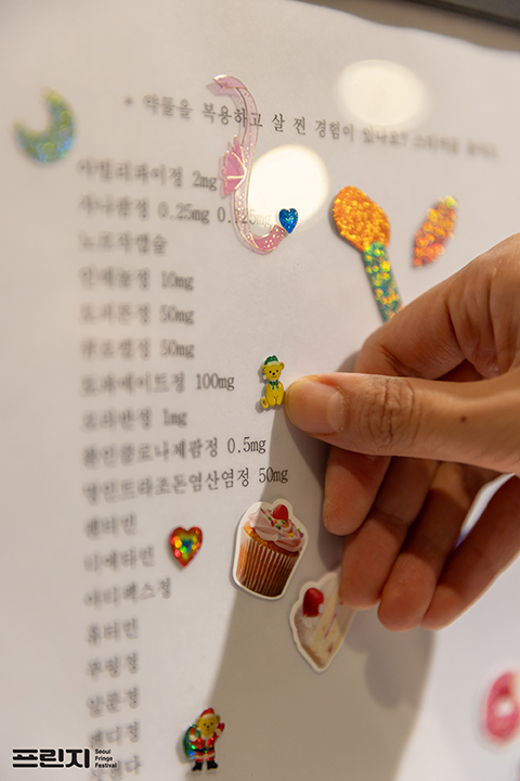 <녹로수>의 전시 사진. ‘약물을 복용하고 살 찐 경험이 있나요?’라는 질문 아래 다양한 약물의 이름이 적힌 종이가 보인다. 
            누군가의 손이 한 약물의 이름 옆에 곰돌이 스티커를 붙인다. 
            카메라의 초점이 맞지 않아 약물의 이름은 정확히 보이지 않고, 종이곳곳에는 다양한 크기와 모양의 반짝거리는 스티커들이 이미 붙어있다. 