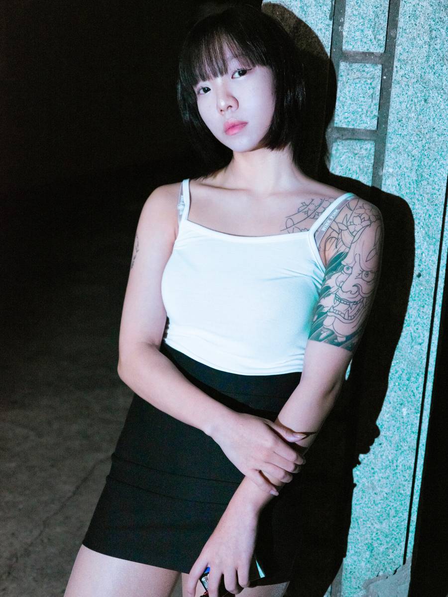 단발머리를 한 인물이 강한 조명을 받으며 카메라를 응시하고 있다. 왼쪽 어깨와 위쪽 팔에 걸쳐 문신을 새겼다. 흰색 민소매 상의와 검은색 치마를 입고 있으며, 오른손으로 왼쪽 팔뚝을 잡고 있다.