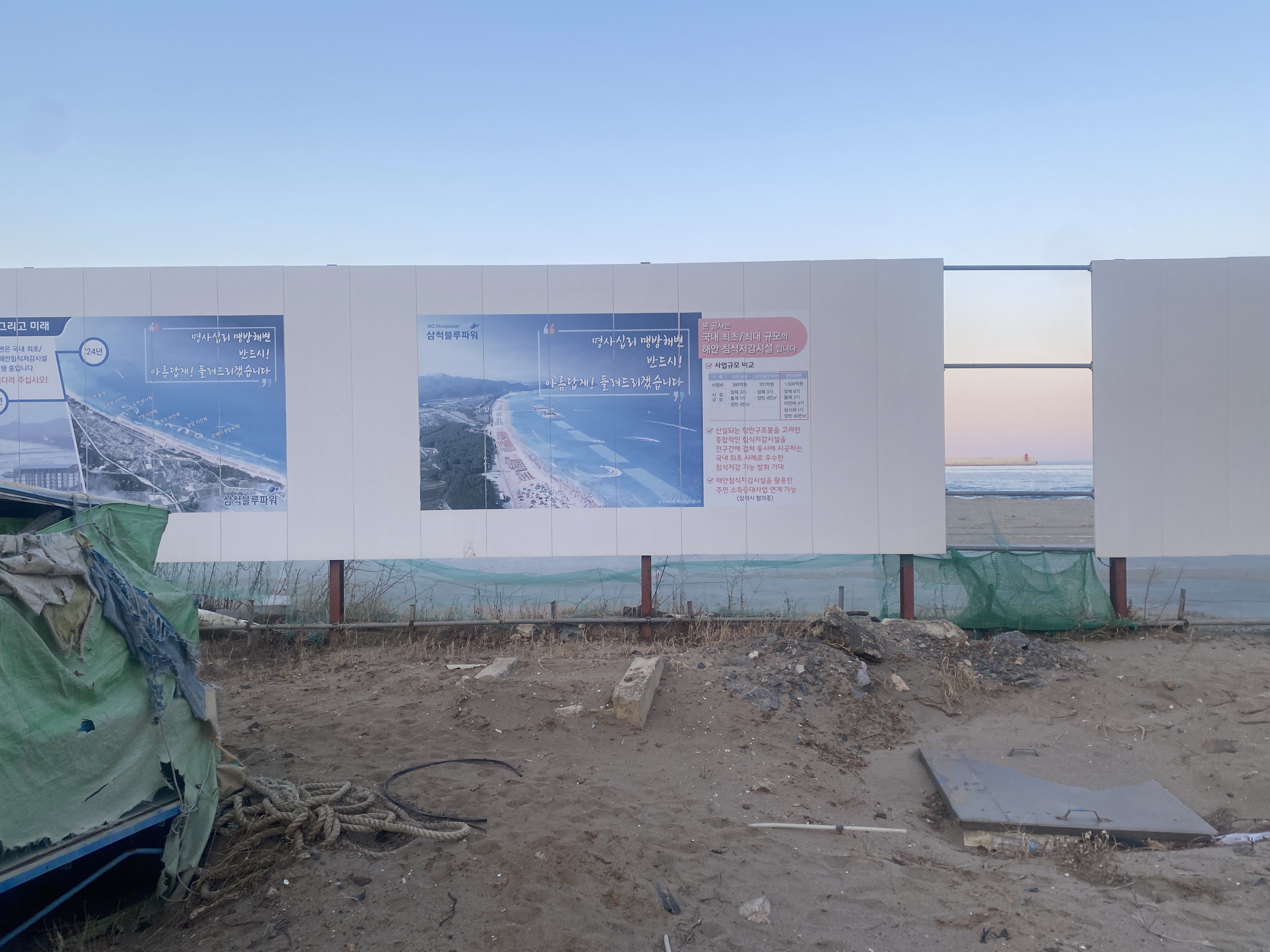 방치된 공사현장처럼 보이는 삼척 맹방해변의 사진. 공사현장의 가림막 뒤로 바다가 보이고, 버려진 밧줄과 그물망, 고무 등이 곳곳에 있다. 공사현장의 가림막에는 “명사십리 맹방해변 반드시! 아름답게! 돌려드리겠습니다”라고 쓰여 있다.