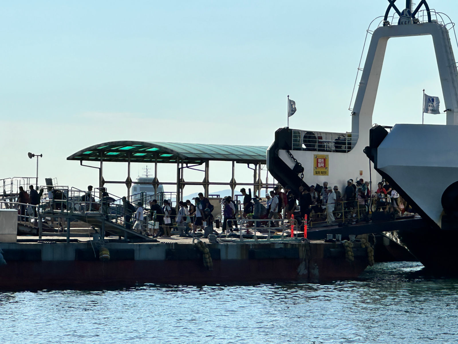 선착장에 배가 도착해 수십명의 사람이 하선하고 있다. 대부분 반팔 차림이며 하늘은 구름 한점 없이 맑다. 