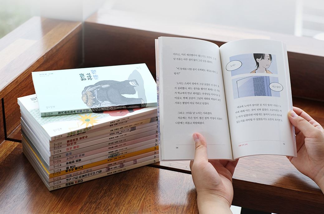 갈색 테이블 위에 놓인 책들과 그 책을 읽고 있는 장면을 찍은 사진이다. 사진의 왼쪽에는 갈색 테이블 위에 쌓여 있는 열네 권의 책이 찍혀 있다. 맨 위에 놓인 책의 제목은 ‘청기와 주유소 씨름 기담’이다. 사진의 오른쪽에는 누군가 양손으로 펼친 책의 내부가 보인다. 펼쳐진 책의 왼쪽 면에는 글이, 오른쪽 면에 글과 그림이 있다. 