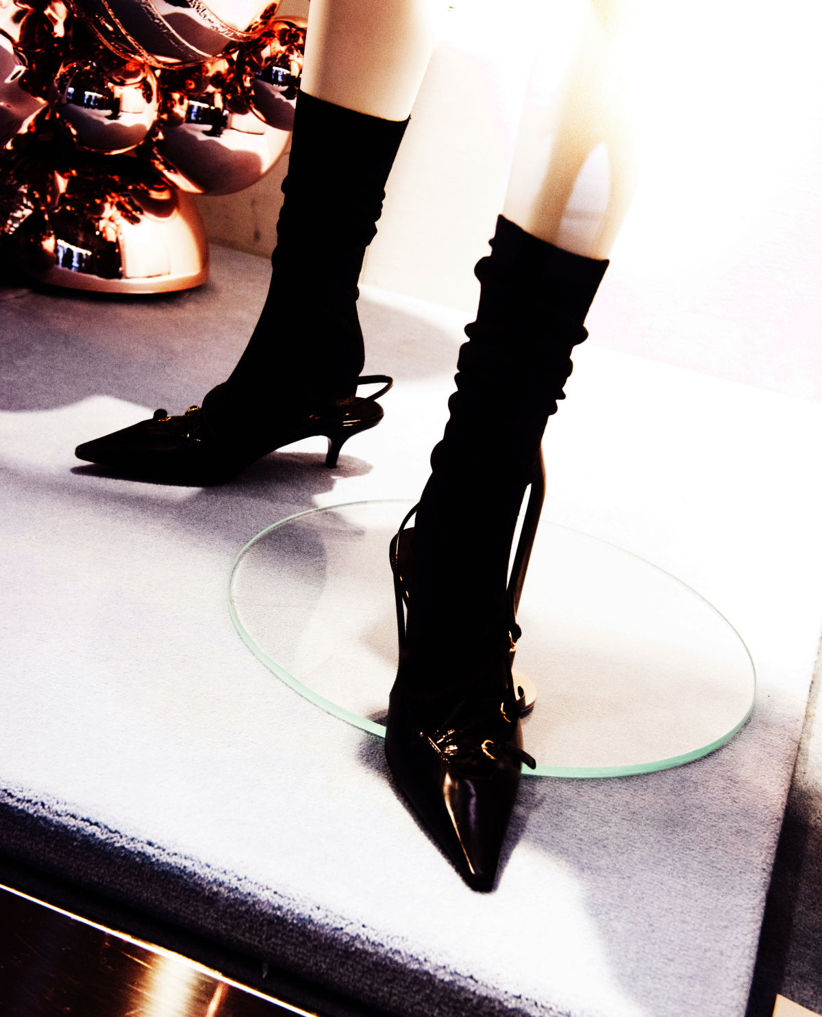 진열대 위, 마네킹의 발목 부분을 찍은 사진. 마네킹에는 검은 양말과 반짝이는 표면과 얇은 끈 디자인이 돋보이는 슬링백힐이 신겨져 있다. 